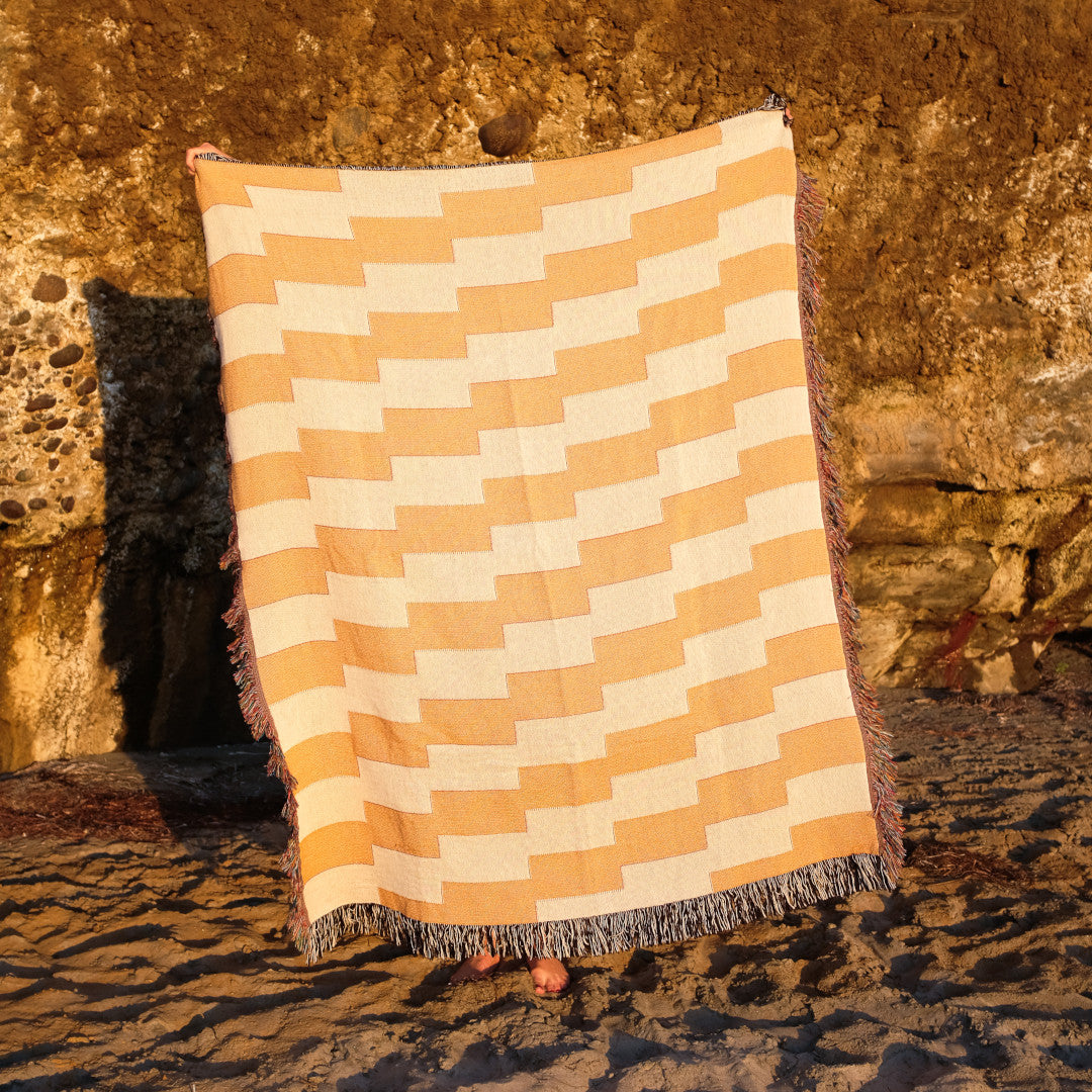 Product image of orange and white striped woven throw blanket titled Amalfi Club at the beach in front of a cliff.