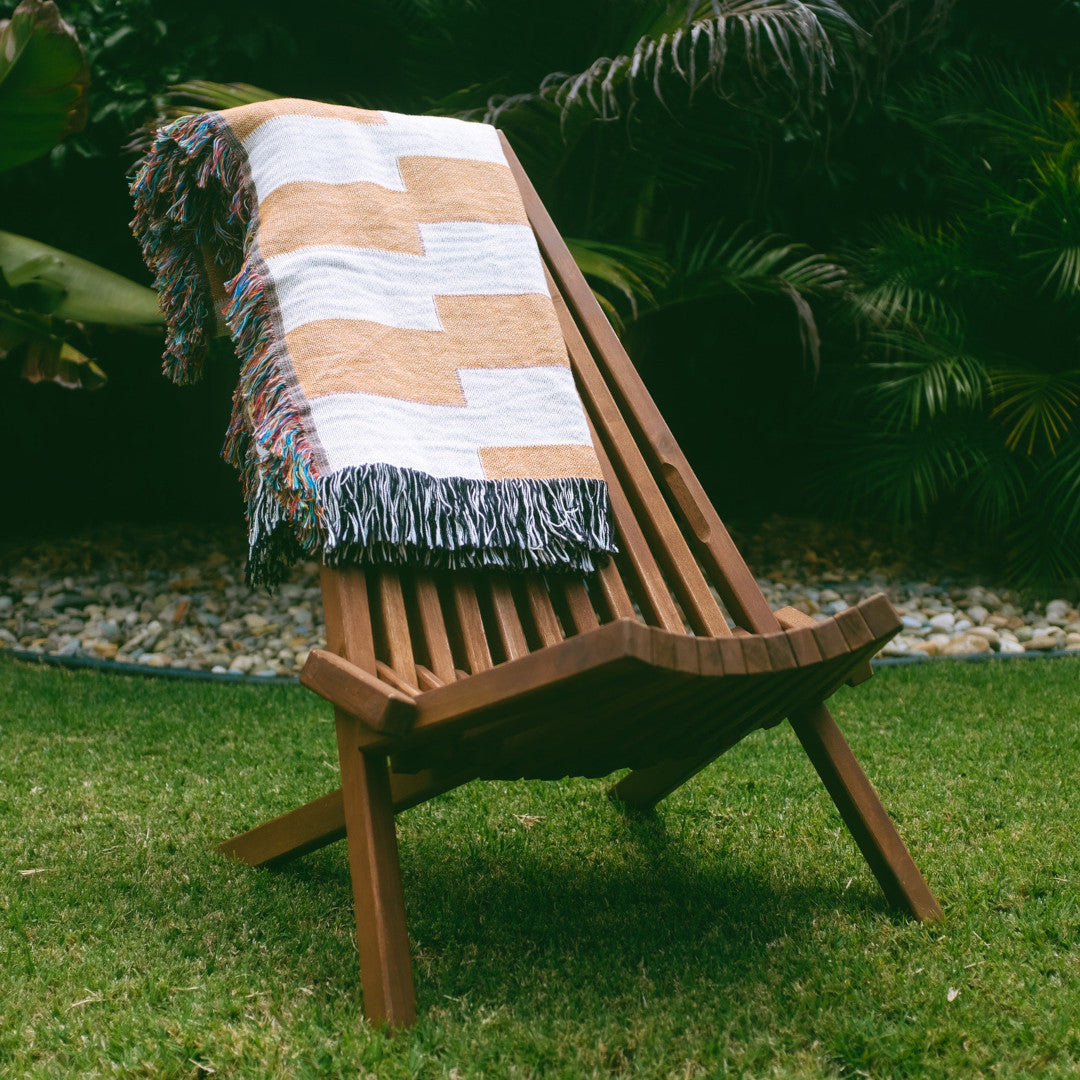 The Amalfi Club orange and white striped woven throw blanket laid on a brown wooden chair outside on grass..