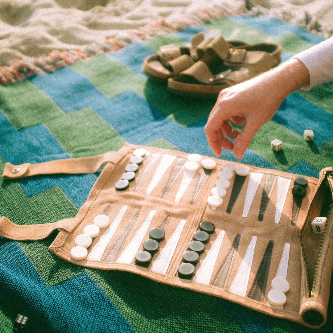 The Capri Club green and blue striped woven throw blanket laid on sand at the beach with a backgammon game sitting on top of blanket.