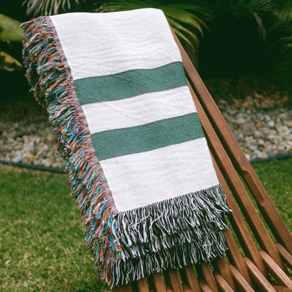 The Fletcher Stripes white and green striped woven throw blanket laid on a brown wooden chair outside on grass..