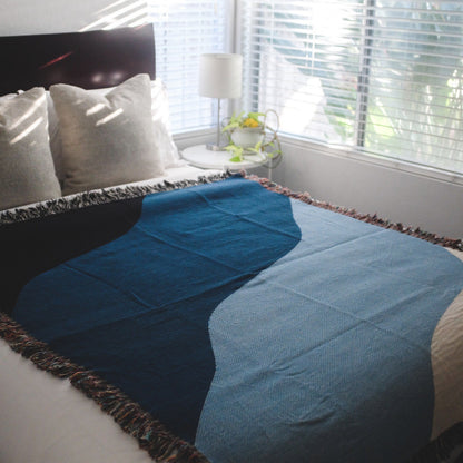 The Shores white, light blue, blue, and black woven throw blanket laid on a white bed in a bedroom next to a window.