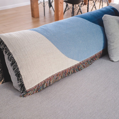 The Shores white, light blue, blue, and black woven throw blanket laid on a light grey couch.