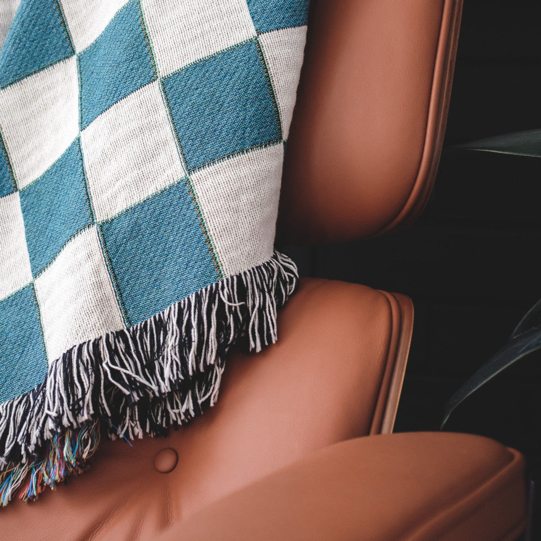 The Solana Checkered teal and white checkered woven throw blanket laid on a brown leather chair.