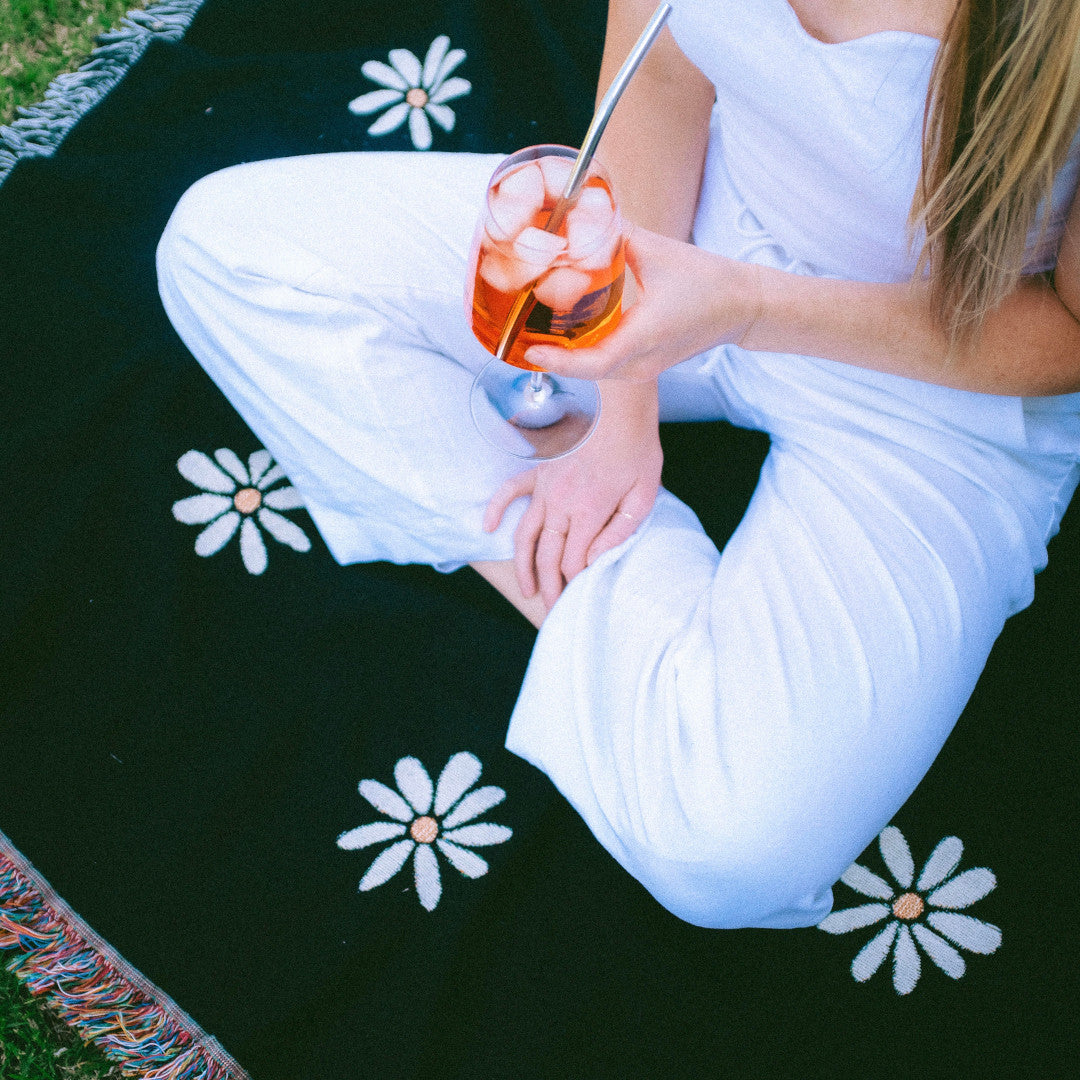 The Supebloom black with white flowers woven throw blanket laid on green grass and a girl dressed in white with orange drink sitting on top of blanket.